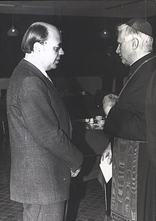 Schwarzweißfotografie von zwei Männern im Profil, die sich gegenüberstehen. Der linke Mann trägt einen Anzug sowie eine Brille und hat eine Halbglatze. Der rechte Mann trägt die Kleidung eines Kardinals und hält einen Zettel in der rechten Hand.