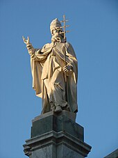 Farbige Untersicht von einer hellen Marmorskulptur. Die Figur trägt ein Bischofsgewand und hält in der linken Hand ein Papstkreuz. Der rechte angewinkelte Arm zeigt in Richtung Himmel.