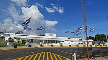 A photograph of the entrance of Hospital El Salvador with several Salvadoran flags waving on flag poles in front of the hospital