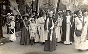 Image of a group of women mostly in light coloured dresses with sashes. Charlotte Despard leads the group holding a cane and a bouquet of flowers and behind her other women hold a Women's Freedom League banner and banners with the names of women. One woman wears a gown and mortar board. A child can also be seen in the front row. A woman stands to the left offering a copy of the newspaper "Votes for Women" to a man, while other people look on. Another woman's sash reads "Women's Freedom League"