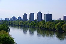 View along the Sava from the embankment opposite to Trnje. The river occupies the bottom of the picture, flanked by rows of deciduous trees. A number of large buildings, mostly brutalist in style, is located behind the trees.