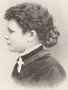 A black and white photograph of a white woman with dark, medium length hair, in profile, wearing a dark dress with a white cross on her lapel.