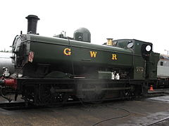 A pannier tank locomotive is standing over an inspection pit. The locomotive is mainly painted green above the running plate, although the chimney is black and the safety valve cover is polished brass. The letters "G W R" are shown in yellow with red shading on the side of the pannier tank. The front buffer beam is painted red.