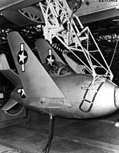 Black-and-white photograph of small potato-shaped fighter aircraft hoisted by a lifting mechanism underneath a large bomber.