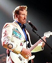 A man in a white jacket and blue shirt plays a white guitar.