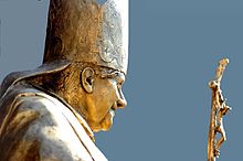 Seitliche Nahaufnahme in Farbe von einer Bronzebüste des Papstes mit liturgischer Kopfbedeckung. Auf der rechten Seite ist der obere Teil des Kreuzstabes zu sehen.
