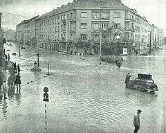 A flooded street corner seen from above. About a dozen people are standing in shallower water. Several others are wading across the intersection. One car is stranded in the water.