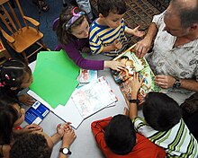 A man sits with a group of children, and points to an image in a picture book.