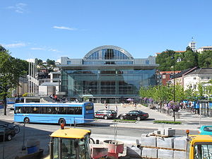 The front of Arendal Town Hall, primarily made of glass. A road in front of the Hall has parked cars and a bus.