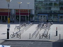 View from high elevation of four ranks of dozens of U-racks set in concrete in front of a glass-fronted building, with disabled car parking spaces in the foreground.