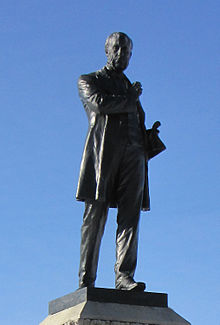 A dark-grey statue of a man standing upright with his left foot forward and his right arm crossed in front of his body