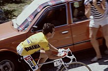 Photograph of Hinault, taken from the side and from further distance, wearing a yellow jersey while riding up a mountain