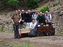 A group of men posing with two wagons. Bottles of wine and various foods are visible in the wagons.