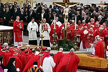 Farbiger Blick auf einen Altar mit Jesus am Kreuz und einem schlichten Sarg, um die eine Gruppe von kirchlichen Würdenträgern in roter und weißer Kleidung steht. Im Hintergrund sind dunkel gekleidete Männer und Frauen zu sehen.