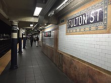 The northbound platform of the Lexington Avenue Line at Fulton Street. A mosaic plaque with the letters "Fulton St" is placed on a wall to the right.