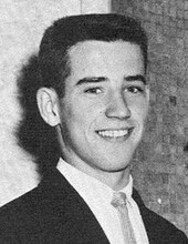 Close-up black-and-white portrait photo of a young Biden smiling and looking directly at the camera, wearing a suit and tie