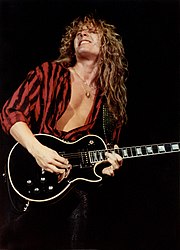 A man with long shaggy hair showing his chest with an unbuttoned shirt and playing guitar.