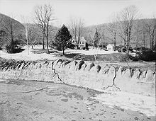 Black and white photograph of a road lined by a low, cracked stone wall with a house and wooded mountains in the background.