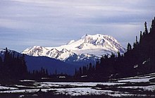 A snow-covered mountain looming over a smaller snow-covered mountain with trees in the foreground