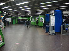A large space covered with dark floor tiles and strip lighting. Ticket machines in the distance and posters on display boards