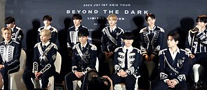 Eleven members of JO1 sitting in two rows in front of a backdrop of JO1 1st Asia Tour Beyond the Dark Limited Edition