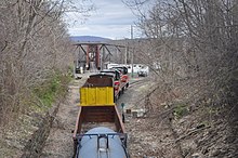 A P&W train is seen from a bridge which crosses over the track it is on. It is moving away from the viewer, and lead by two diesel locomotives, followed by several assorted freight cars. A railroad swing bridge can be seen in the distance.