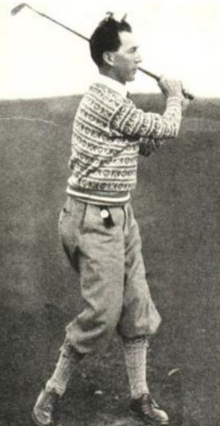 A white man with dark hair, in a golf swing pose, wearing a knitted sweater and short trousers
