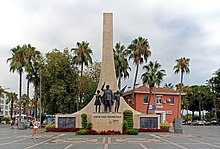 A tall sweeping stone triangle projects skyward behind the statues of a man and two children in bronze on a smaller podium. Around the base are placed several wreaths with logos. Palm trees surround the scene.