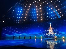 Photograph of the stage lit in blue, with at the back a wide LED-screen with the Junior Eurovision 2021 logo with the text "Imagine"; above the stage are numerous blue and yellow lights