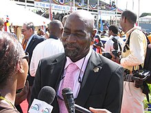 A bald black man in a suit, white shirt and pink tie, smiling and being interviewed by a female in glasses