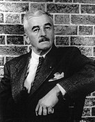 A photograph of writer William Faulkner dressed in a suit and seated in a chair.