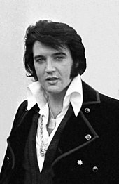 Posed photo of Elvis, shoulders, and chest, wearing a shirt with a large, white collar and a coat with big lapels, medallions around his neck.
