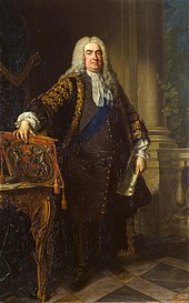In a full-length portrait, an overweight middle-aged man stands, legs slightly apart, with his right hand resting on the top of a coat of arms, leant on the top of an ornate table. In his left hand he holds a large white document. He is dressed in expensive 18th-century clothing, with a blue sash over his left shoulder, and a long grey wig. Behind him, a large pale column of stone rises from a tiled floor. A balustrade connects to the column, and a small green plant is visible.