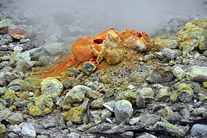 A rocky surface with a mound centrally in the image. The mound has holes in the top. Surrounding rocks have been stained orange and yellow. There is smoke or steam rising from the top.