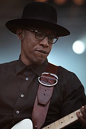 A black man in a black button-down shirt, black-frame eyeglasses, and a black fedora playing an electric guitar and biting his lip