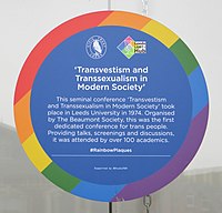 A circular commemorative plaque with a thick rainbow border. The logos of the Leeds Civic Trust and the Leeds LGBT+ Hub are visible near the top. "'Transvestism and Transsexualism in Modern Society'" is written below. Below that is the following text in smaller print: "The seminal conference 'Transvestism and Transsexualism in Modern Society' took place in Leeds University in 1974. Organized by The Beaumont Society, this was the first dedicated conference for trans people. Providing talks, screenings and discussions, it was attended by over 100 academics." The two lines below, in smaller print, read "#RainbowPlaques" and "Supported by @StudioTDH".