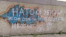 White spray-painted text on a tan wall that reads "НАТО СЕ УБИЈЦИ ЈАС СУМ СПАС ЗА МАКЕДОНИЈА#БОЈКОТ" which translates to "NATO are killers. I am for the salvation of Macedonia.#Boycott."