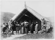 Photograph of a group of Maori between 1881 and 1886, in front of the Hinemihi meeting house at Te Wairoa, Lake Tarawera, New Zealand. Āporo Te Wharekāniwha is holding a club aloft, and died of illness in Rotorua's Sanatorium Hospital on 23 May 1886.