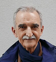 senior White man with short grey hair and moustache, wearing a blue jacket and scarf, smiling at camera