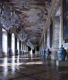 A view down the Ahnengalerie. The walls, made of scagliola, are lined with portraits of the rulers of Württemberg, their arms, and an urn. Above the gallery is a massive fresco, featuring several Classical figures, glorifying the reign of Eberhard Louis.
