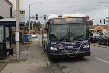 An articulated transit bus stopped at the curb on a road, next to a sidewalk and overhead shelter with a bench and garbage can. A utility pole is furnished with a sign listing various bus routes.