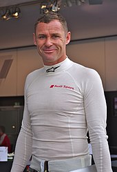 Tom Kristensen smiling at the camera and wearing white racing overalls