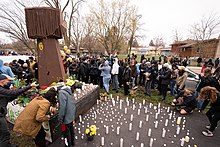 A group of people gather on a sidewalk along a street. There are candles, flowers, and a large, wooden fist sculpture.