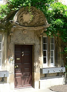 A large decorated shell-like stone canopy, with a shield in the centre and carved foliage around; beneath it, a wooden door with brass handle and knocker, set into a stone building; foliage grows above and around the canopy