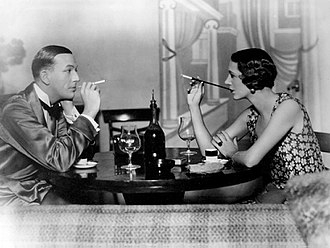 young white man, clean shaven, facing a young white woman with bob cut hair across a small round table set with wine glasses and demitasse cups; both are smoking using cigarette holder