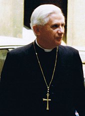 Verschwommene Farbfotografie von Ratzinger in schwarzer Priesterkleidung mit einem goldenen Kreuz als Halskette.