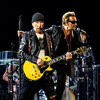 The Edge and Bono clothed in leather jackets, as the Edge holds a guitar vertically. A large dangling light bulb hangs between them.