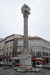 Color photograph of an ancient stone column set in front of a modern building and parked cars. The bottom and top of the column are engraved, and several metal bands placed at regular intervals encircle the central section of the column.