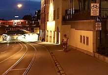 Eine lange Bahnunterführung in Augsburg mit leichtem Gefälle, glänzende Meterspur-Straßenbahnschienen mit etwa 1,50 m Abstand vom Bordstein, Überholverbot (durchgezogene weiße Linie) für Autos in der beleuchteten Unterführung nahe dem Hauptbahnhof mit einem rot-weißen Zug oberhalb der leicht gebogenen Tunnelüberwölbung. Eine Fahrradfahrerin (vermutlich) auf dem etwa 2,50 Meter breiten Gehweg rechts, ein Bettler sitzt ein wenig weiter unten am Rand an der hohen Hauswand. Verkehrsschild Fussgänger mit Zusatzschild Fahrradfahren frei und Hinweistafel „Radfahren auf der Fahrbahn erlaubt“. Links auf der gegenüberliegenden Straßenseite trennt keine Wand mehr den Bürgersteig von der aufwärtsführenden Fahrbahn ab. Blaue Stunde am Abend mit Gebäude einer Brauerei rechts im Bild.