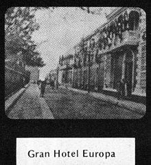 A fuzzy black-and-white still frame showing a street scene with a hotel on the right and a tall fence on the left. A man appears to be stepping into the road. The still frame is surrounded by a black border frame, which is annotated below with the printed words "Gran Hotel Europa".